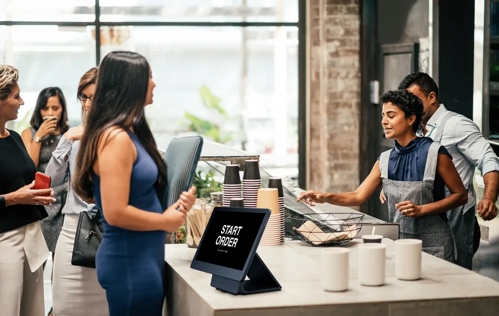 Guests placing orders at a Shift4 Dine self-order kiosk in a busy cafe